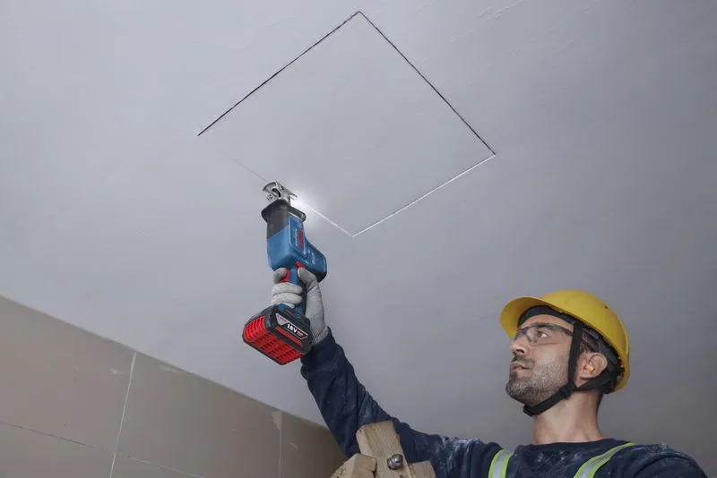 Una persona que usa equipo de seguridad corta un panel de techo con una sierra alternativa.