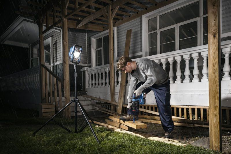 Una persona que usa equipo de seguridad perfora madera en un porche, iluminado por una luz de trabajo inalámbrica.