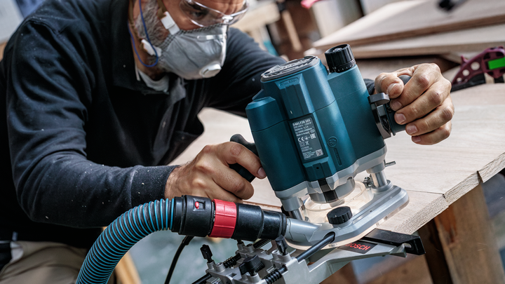 Una persona que usa equipo de seguridad utiliza una fresadora eléctrica para dar forma al borde de una tabla de madera.