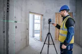 Una persona que lleva equipo de seguridad maneja una herramienta de nivelación láser sobre un trípode en una habitación de hormigón.
