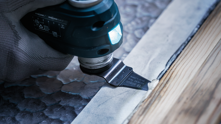 Una persona que usa equipo de seguridad utiliza una herramienta múltiple para cortar material junto a molduras de madera.