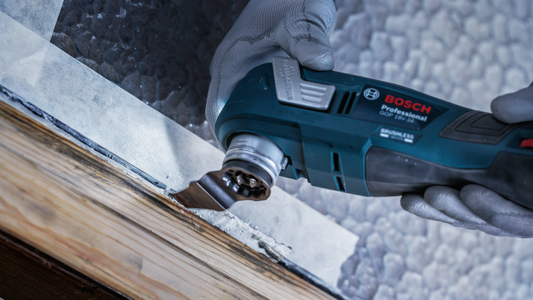 Una persona que usa equipo de seguridad utiliza una herramienta múltiple para raspar la pintura de un marco de ventana de madera.