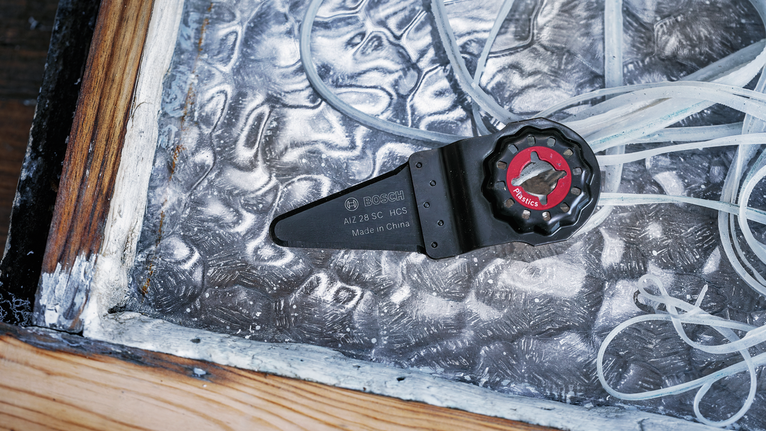 El accesorio de hoja de herramienta oscilante descansa sobre una lámina de metal texturizada cerca de un marco de madera.