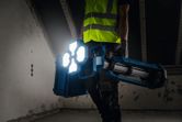 Una persona con equipo de seguridad porta una luz de obra inalámbrica en una habitación con poca luz.
