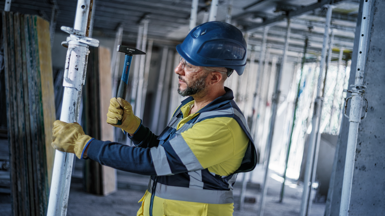 Una persona que lleva equipo de seguridad utiliza un martillo para ajustar un andamio en una obra de construcción.