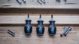 Tres destornilladores dispuestos sobre un trozo de madera junto a tornillos y pernos dispersos.