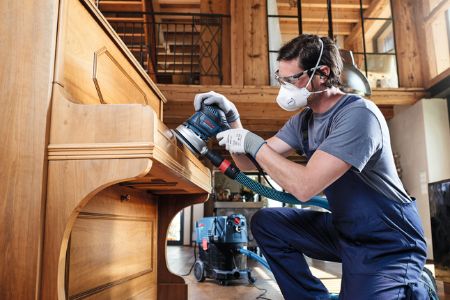Una persona que usa equipo de seguridad lija un piano de madera con una lijadora eléctrica.