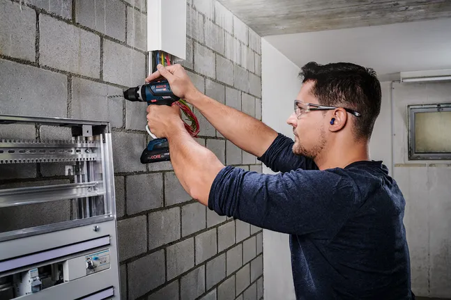 Una persona, equipada con equipo de seguridad, taladra una pared de bloques de hormigón cerca del cableado eléctrico.