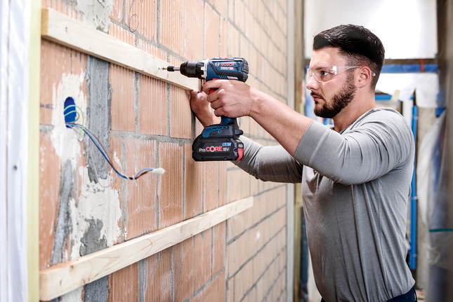 Una persona que lleva equipo de seguridad utiliza un taladro eléctrico para fijar madera a una pared de ladrillos.
