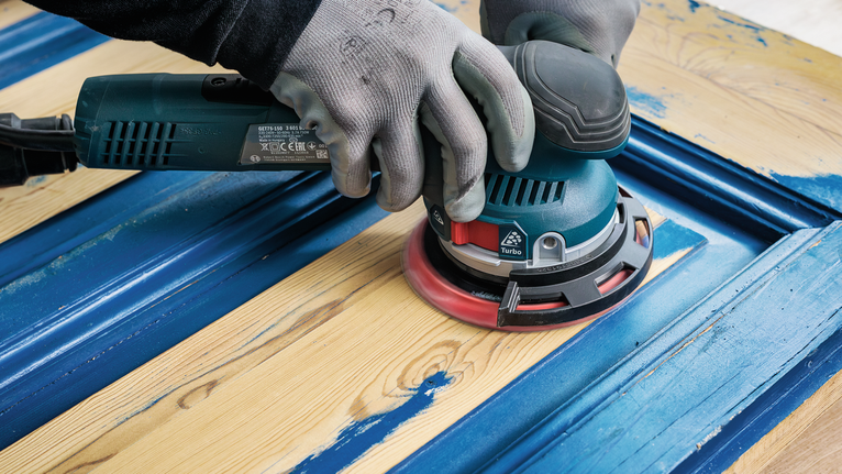 Una persona que usa equipo de seguridad lija pintura azul de una puerta de madera.