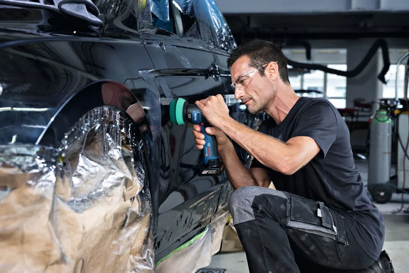 Una persona que usa equipo de seguridad pule la puerta de un automóvil con una herramienta inalámbrica.