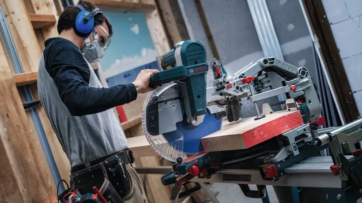 Una persona que lleva equipo de seguridad corta una tabla de madera con una sierra ingletadora deslizante.