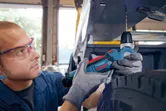 A person wearing safety equipment uses a cordless angle drill to bore metal.