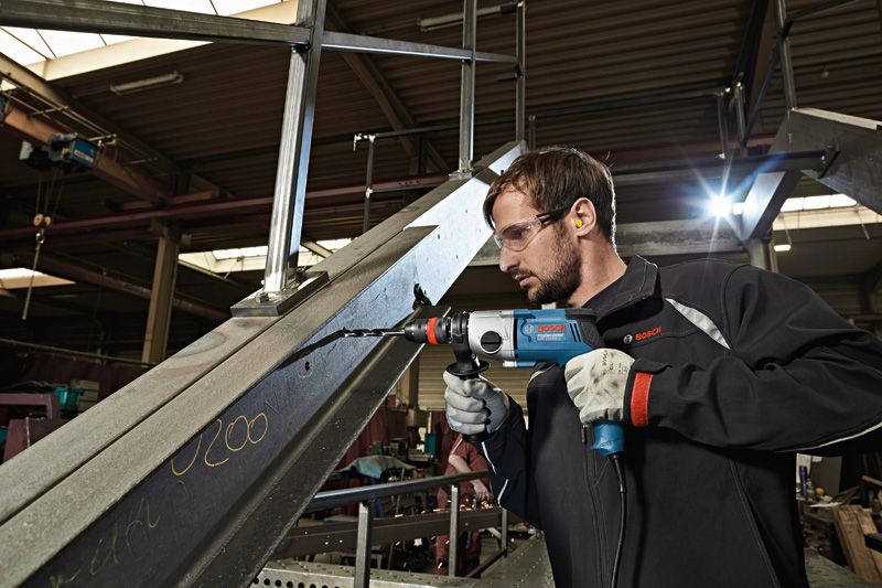 A person wearing safety equipment uses a drill to make a hole in a metal beam.