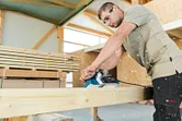 A person wearing safety equipment uses a planer to smooth a wooden beam.