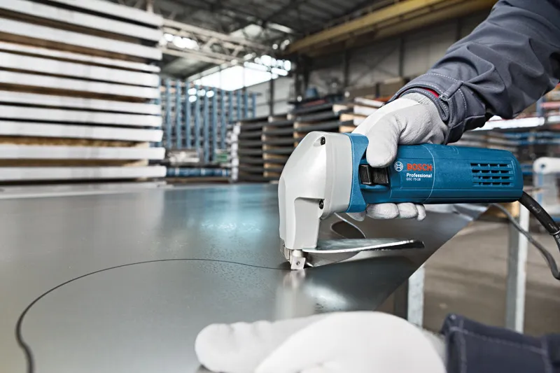 A person wearing safety equipment uses an electric shear to cut sheet metal.