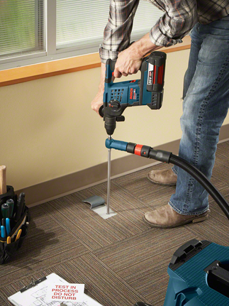 Worker drills into an office floor with a rotary hammer, wearing safety equipment.