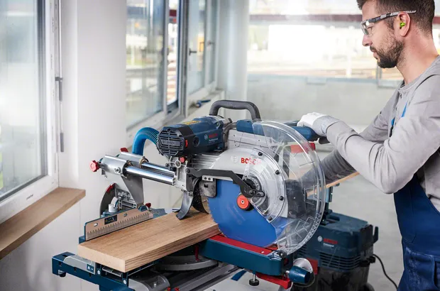 Person wearing safety equipment uses a sliding miter saw to cut a wooden plank.