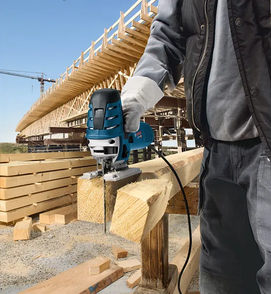A person wearing safety equipment uses a jigsaw to cut a wooden beam at a construction site.
