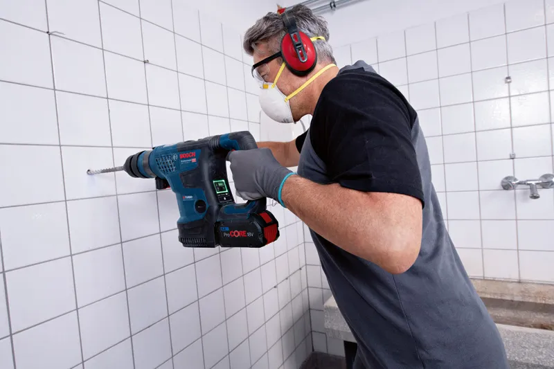A person wearing safety equipment drills into a tiled wall with a cordless rotary hammer.