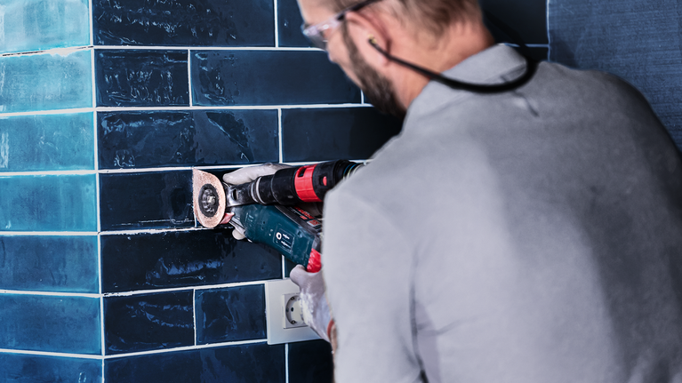A person wearing safety equipment grinds dark wall tiles with a power tool.