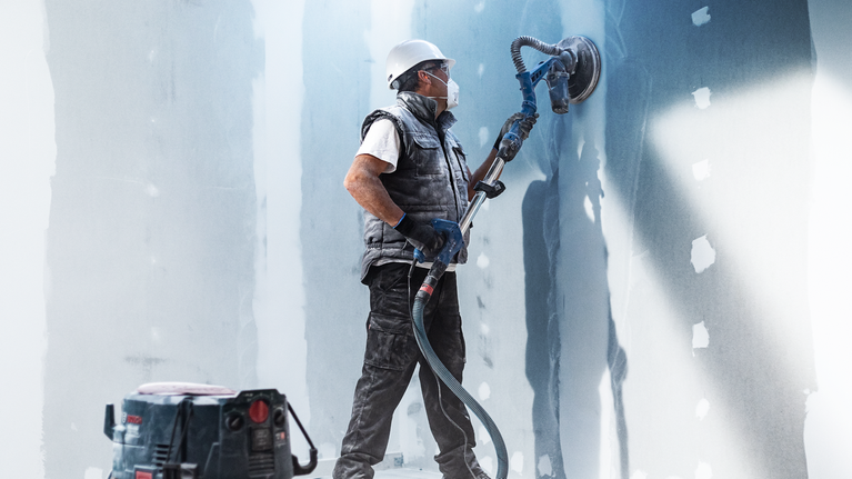 A person wearing safety equipment sands drywall with a long-handled power tool.