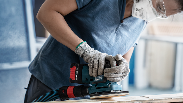 Person wearing safety equipment sands wood with a power sander.