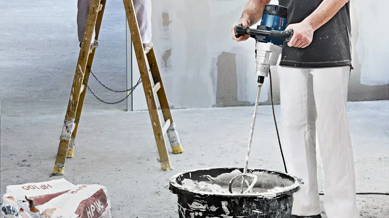 A person wearing safety equipment uses a stirring mechanism to mix material in a bucket.
