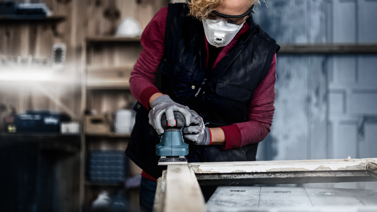 Person wearing safety equipment sands wood with a power tool in a workshop.