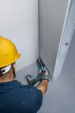A person wearing safety equipment fastens drywall with a cordless screwdriver.