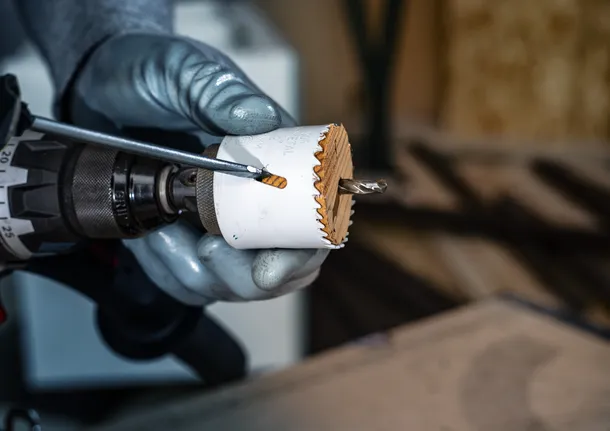 Person wearing safety equipment uses a drill with a hole saw to cut through wood.