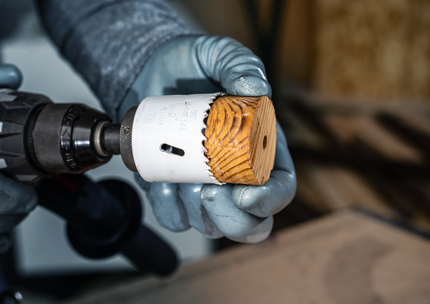 Person wearing safety equipment holds a drill with a hole saw bit containing a wood core.