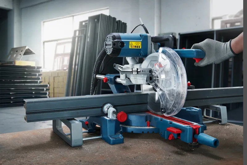 A person wearing safety equipment operates a miter saw to cut a metal bar.