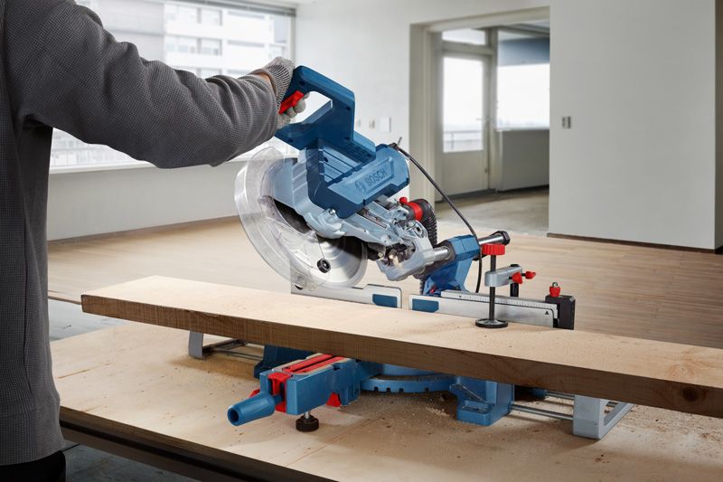 A person wearing safety equipment cuts a wooden plank using a miter saw.