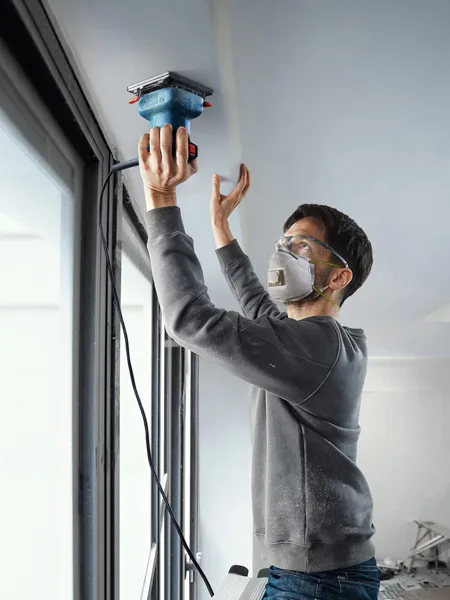 A person wearing safety equipment sands a ceiling edge with an orbital sander.