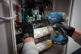 Person wearing safety equipment uses a cordless drill on an electrical control panel.