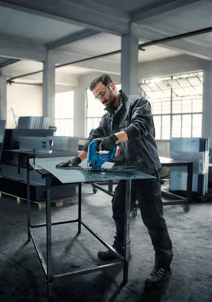 A person wearing safety equipment uses a jigsaw to cut sheet metal in a workshop.