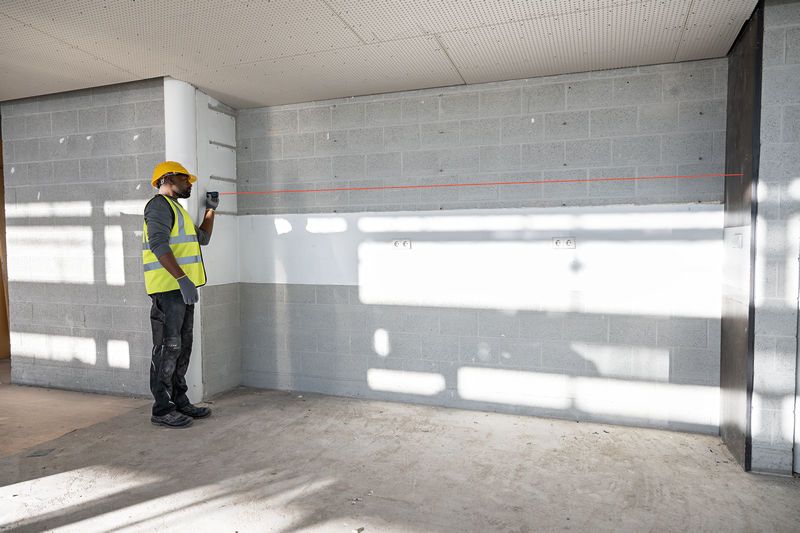 A person wearing safety equipment uses a laser measure to mark a straight line on a wall.