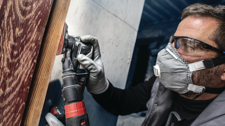 Person wearing safety equipment sands a wooden surface with a power tool.
