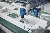 A person wearing safety equipment uses a cordless drill to assemble electronic components.
