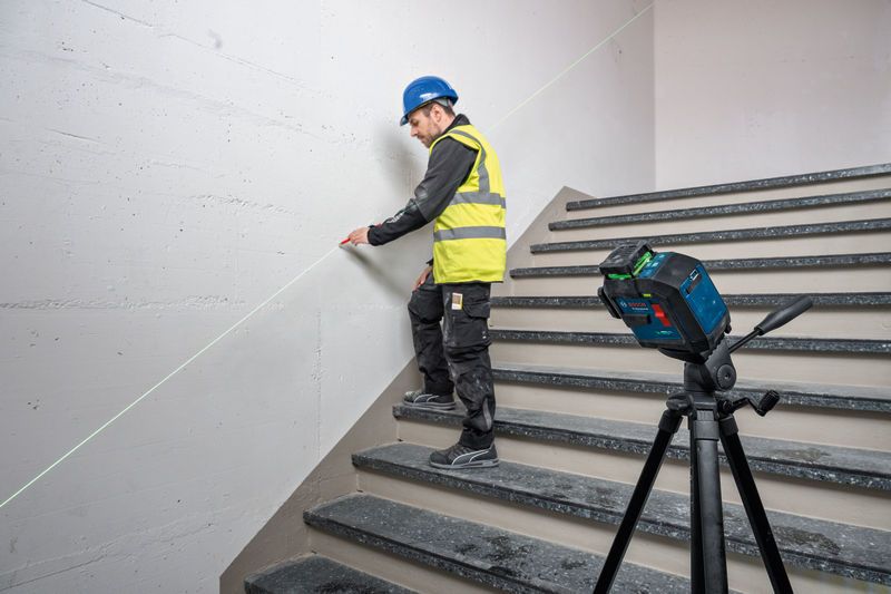 A person wearing safety equipment marks a wall using a laser leveling tool on a tripod.