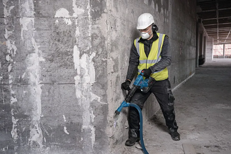 A person wearing safety equipment breaks concrete with a cordless demolition hammer.