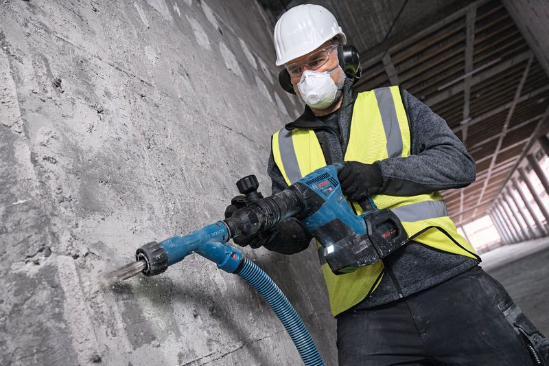 A person wearing safety equipment uses a cordless demolition hammer on a concrete wall.