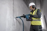 A person wearing safety equipment uses a cordless demolition hammer to break a concrete wall.