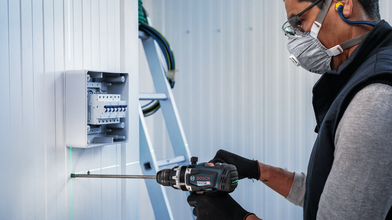 Person wearing safety equipment drills into a wall near electrical casing.