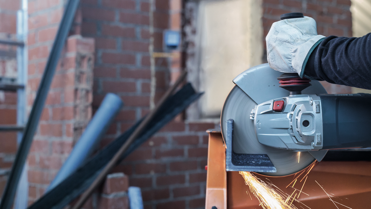 A person wearing safety equipment cuts metal with an angle grinder, creating sparks.