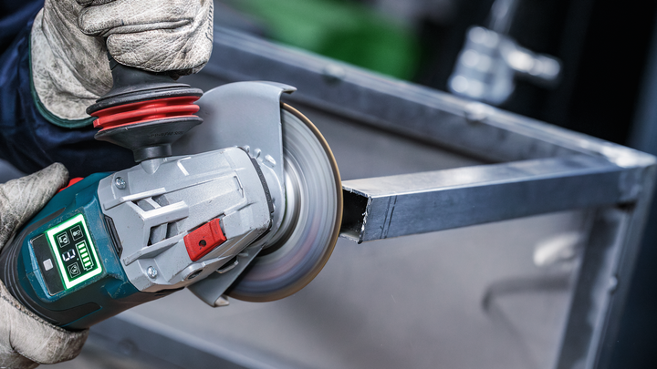 Person wearing safety equipment uses an angle grinder to cut a metal beam.