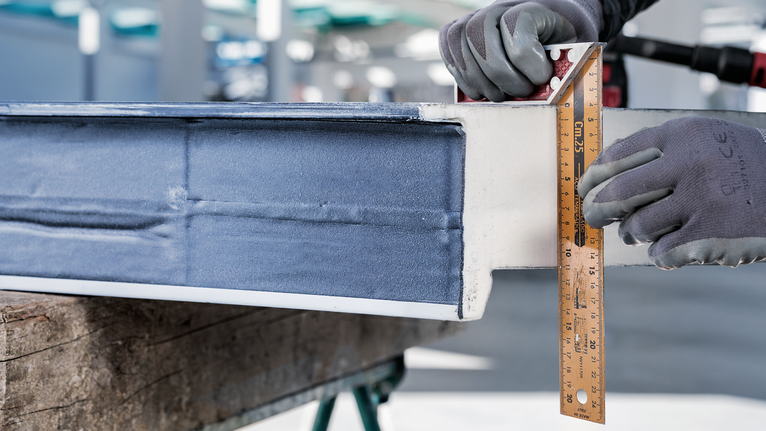 Person wearing safety equipment measures a metal beam with a ruler.