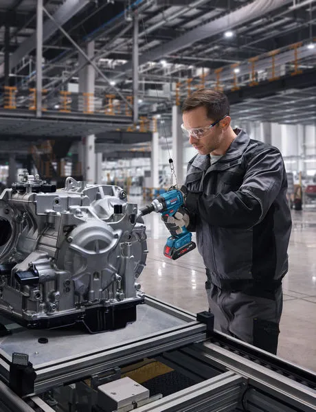 A person wearing safety equipment uses a cordless impact wrench on an engine assembly.