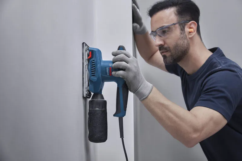 A person wearing safety equipment uses an orbital sander on a white wall.